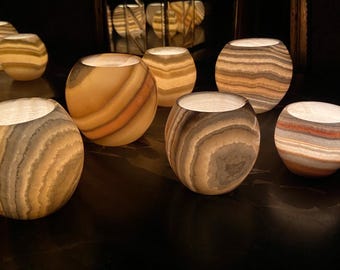 Könnte beinhalten: Eine Gruppe von acht runden, gestreiften Alabaster-Kerzenhaltern mit warmem Schein. Die Kerzenhalter sind in einem Halbkreis auf einer dunklen Oberfläche angeordnet.