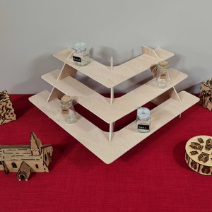May include: A three-tiered wooden display stand with a chevron design. The stand is made of light wood and has four small glass jars with cork tops on the shelves. The stand is on a red surface with other wooden decorative items.