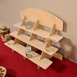 May include: A light-colored wooden display shelf with three tiers, holding several small glass jars with lids. The shelf has a curved top and is set against a neutral wall. A decorative wooden lantern is visible on the left.