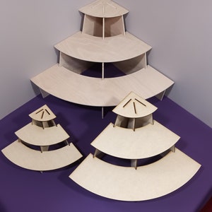 May include: Three unfinished wooden tiered display stands in varying sizes. Each stand has a triangular top and multiple curved shelves. The stands are arranged on a purple surface against a gray wall.