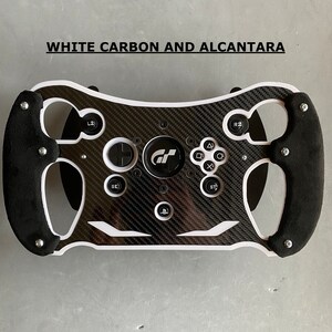 May include: A black and white racing steering wheel with a carbon fiber design and white accents. The steering wheel has a PlayStation button and a GT logo in the center.