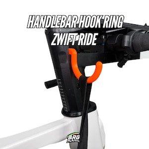 Op de afbeelding: Een zwart stuur met een oranje haakring, met de tekst "HANDLEBAR HOOK RING ZWIFT RIDE" in het wit. Een zwarte riem hangt aan de haak. De basis is wit met een logo.