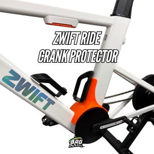 Op de afbeelding: Wit fietsframe met oranje en zwarte crankbeschermer. De tekst "ZWIFT RIDE CRANK PROTECTOR" wordt in het zwart weergegeven. Het Zwift-logo is in blauw en groen.
