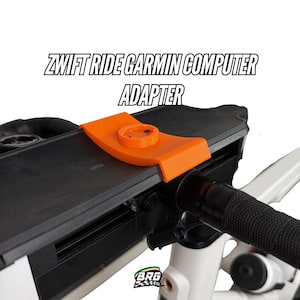 May include: An orange Zwift Ride Garmin computer adapter is mounted on a black device, with the text "ZWIFT RIDE GARMIN COMPUTER ADAPTER" above. The device is attached to a white bicycle handlebar with a black grip.