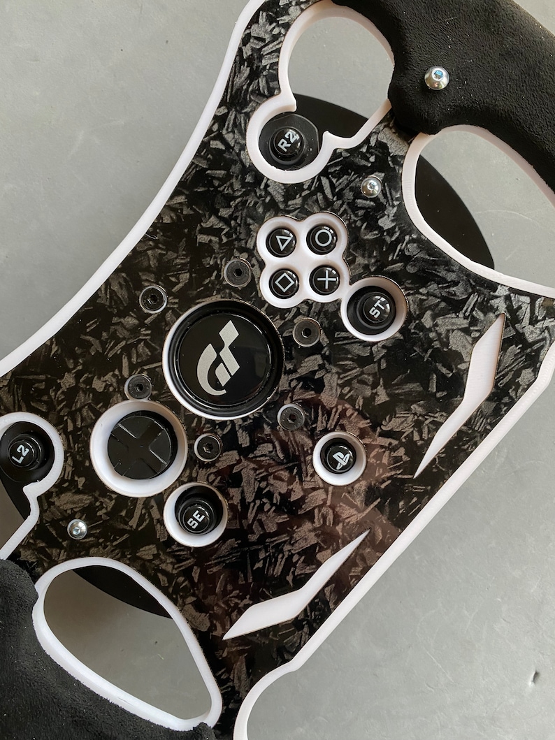 May include: A black and white racing steering wheel with a carbon fiber pattern. The wheel has a variety of buttons, including a PlayStation button, a start button, and buttons labeled L2 and R2. The wheel also has a black and white GT logo in the center.