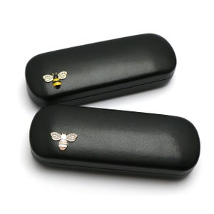 May include: Two black rectangular eyeglass cases with a bee design on the front. The bee is gold with black and yellow stripes and has clear rhinestones.