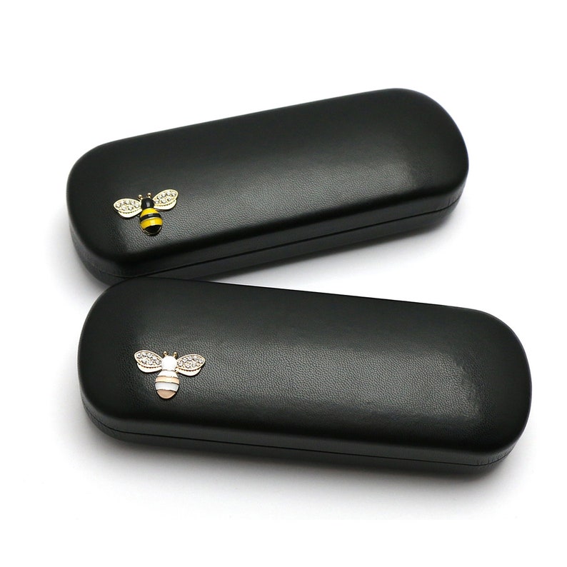 May include: Two black rectangular eyeglass cases with a bee design on the front. The bee is gold with black and yellow stripes and has clear rhinestones.