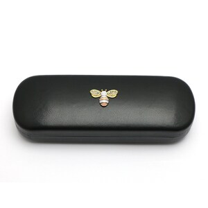 May include: Black leather eyeglasses case with a gold bee design. The bee has white wings and a pink and yellow body.