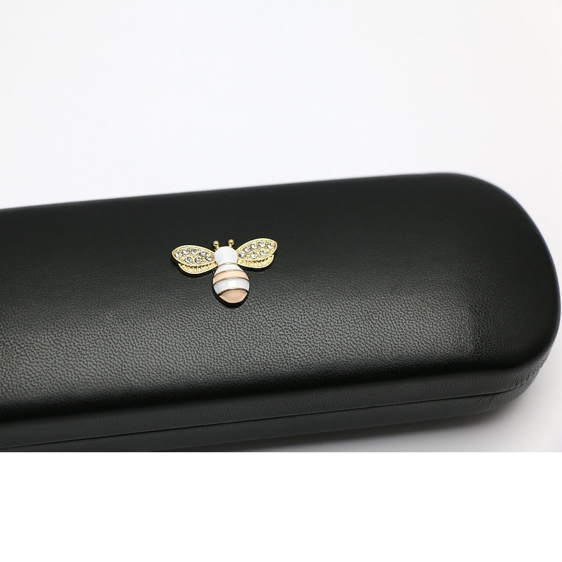 May include: Black leather glasses case with a gold bee design. The bee has white and pink stripes and is decorated with rhinestones.