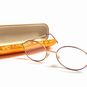 May include: A pair of round, tortoiseshell spectacles with gold metal frames and clear lenses. The spectacles are resting on a gold and orange patterned case.