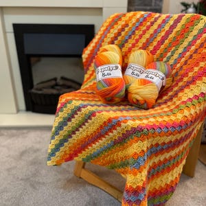 Puede incluir: Una manta de crochet colorida en tonos naranja, amarillo, verde y azul está drapeada sobre una silla de madera. Tres ovillos de lana están encima de la manta, cada uno con la etiqueta "Papatya Batik".