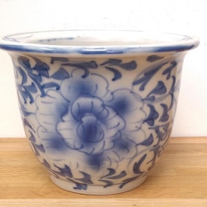 May include: A white ceramic planter featuring a hand-painted blue floral pattern with swirling leaves and vines. The pot has a wide, rounded rim and a tapered base, showcasing a smooth, glossy finish with a classic blue and white colour scheme.