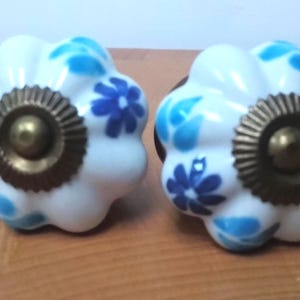 May include: Two white ceramic cabinet knobs with a floral design in shades of blue. Each knob has a brass-coloured base and a rounded, flower-like shape. These decorative knobs are ideal for furniture.