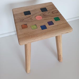 Op de afbeelding: Een houten kruk met vier poten en een vierkant bovenblad. Het bovenblad is versierd met kleurrijke vierkanten en cirkels.