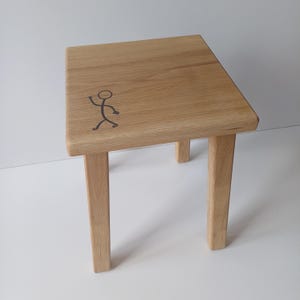Peut inclure: Un petit tabouret en bois avec quatre pieds et un plateau carré. Le dessus du tabouret est décoré d'un simple dessin de bonhomme bâton.
