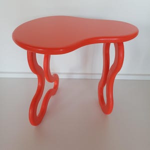May include: A bright orange, freeform shaped side table with a unique, abstract design. The table has a smooth, glossy finish and features two curved legs on each side.
