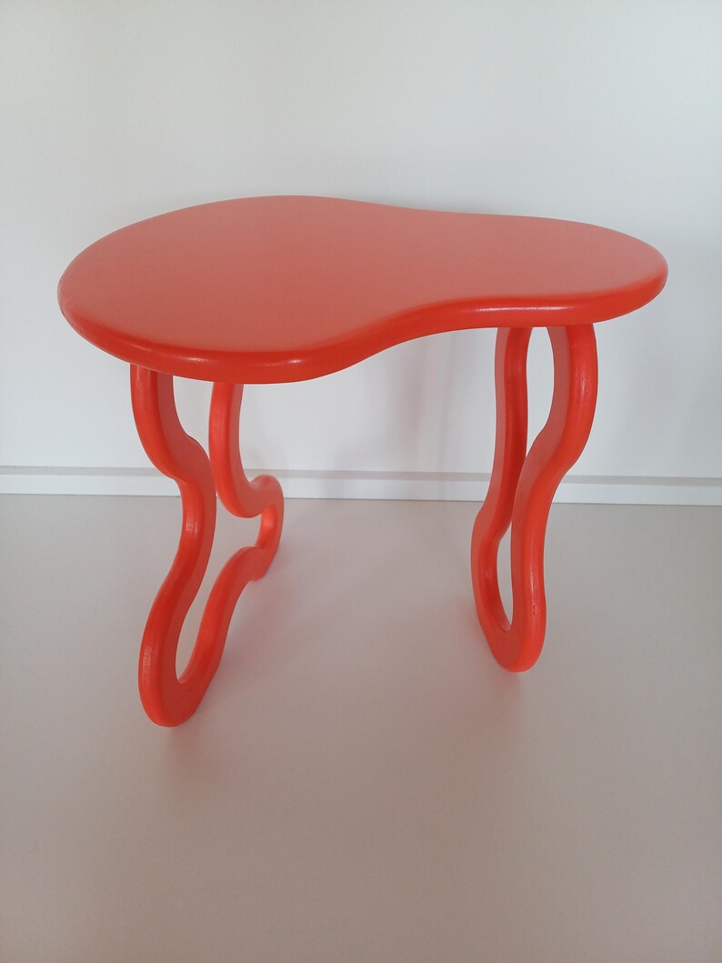 May include: A bright orange, freeform shaped side table with a unique, abstract design. The table has a smooth, glossy finish and features two curved legs on each side.