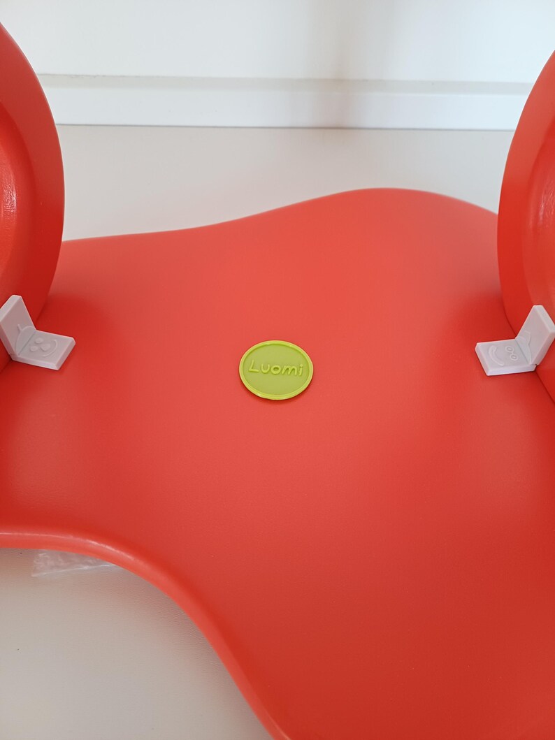 May include: A bright red, wavy, plastic chair with a green round sticker that says "Luomi" on it.