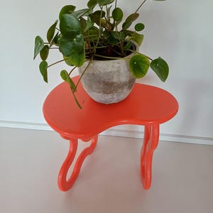 May include: A bright orange plant stand with a unique, wavy design. A potted plant with round, green leaves sits on top of the stand.