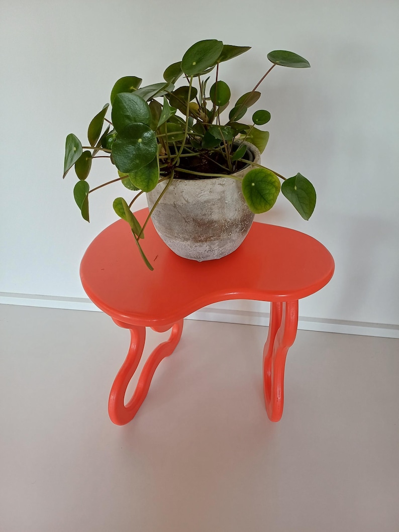 May include: A bright orange plant stand with a unique, wavy design. A potted plant with round, green leaves sits on top of the stand.