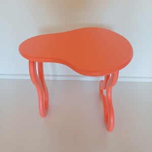 Peut inclure: Une table d'appoint moderne de couleur orange vif. Le plateau, de forme organique, mesure environ 60 cm de long et 40 cm de large. La table est soutenue par trois pieds incurvés, offrant un design élégant et fonctionnel.