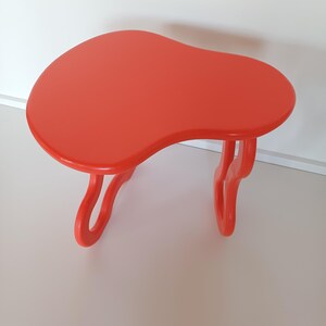 May include: A bright orange, freeform shaped side table with a unique, organic design. The table has two legs that are also shaped like organic forms.