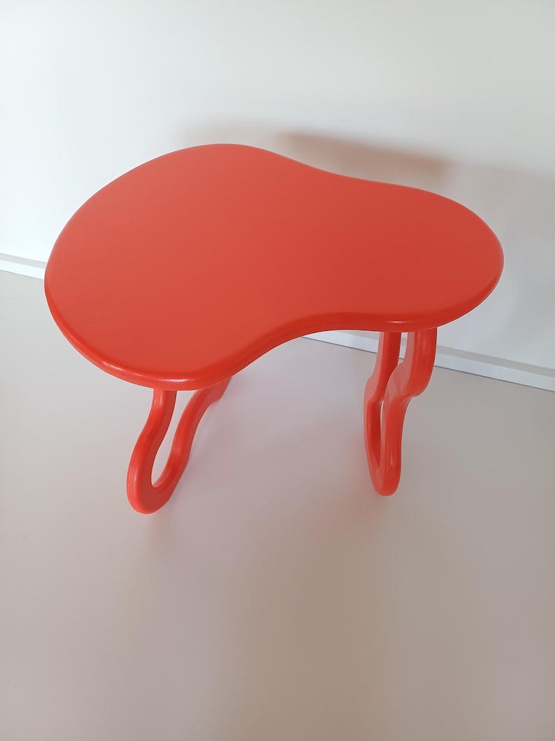 May include: A bright orange, freeform shaped side table with a unique, organic design. The table has two legs that are also shaped like organic forms.