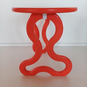 May include: A bright orange, round table with a unique, abstract, wavy base. The table is made of wood and has a smooth, glossy finish.