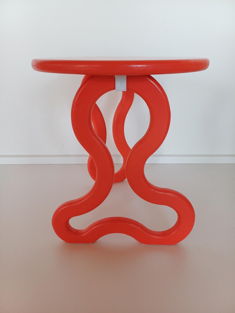 May include: A bright orange, round table with a unique, abstract, wavy base. The table is made of wood and has a smooth, glossy finish.