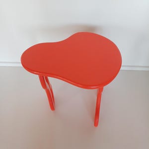May include: A bright orange, freeform shaped side table with a smooth, glossy finish. The table has a unique, organic design with two curved legs.