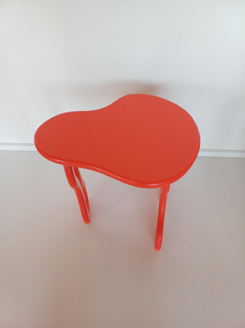 May include: A bright orange, freeform shaped side table with a smooth, glossy finish. The table has a unique, organic design with two curved legs.