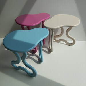 May include: Three abstract, organic-shaped side tables in blue, pink, and white. The tables have a unique, flowing design with rounded edges.