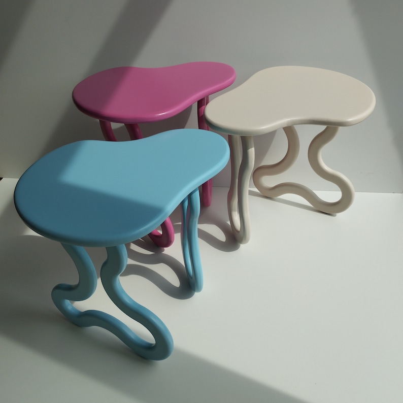May include: Three abstract, organic-shaped side tables in blue, pink, and white. The tables have a unique, flowing design with rounded edges.