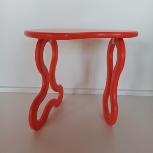 May include: A bright orange, free-form shaped side table with a rounded top and three legs. The legs are shaped like a figure eight.