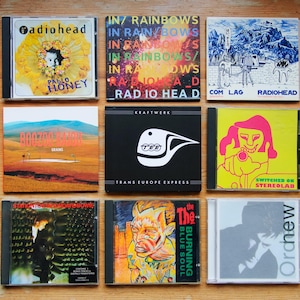 May include: A collection of nine CD cases displayed on a wooden surface. The CDs showcase album artwork from artists such as Radiohead, Kraftwerk, and David Bowie. The album covers feature diverse designs, including colourful illustrations and minimalist graphics.