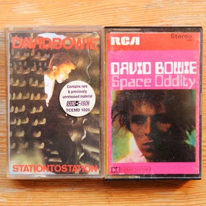 May include: Two vintage cassette tapes featuring David Bowie album covers. One is titled "Station to Station" with a black and white image. The other is "Space Oddity" with a pink and black cover. Both are stereo.