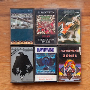 May include: A collection of six cassette tapes featuring the band Hawkwind. The tapes are arranged in two rows of three. The tapes are all black with silver lettering and feature different album covers. The album titles include "Roadhawks", "Stasis", "The UA Years 1971-1975", "The Chronicle of the Black Sword", "Zones", and "Hawkwind - Bonus Tape Track".