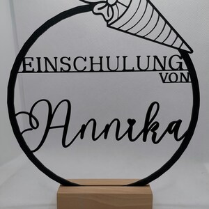 Könnte beinhalten: Schwarzer Metallkreis mit den Worten "EINSCHULUNG VON Annika" und einem dekorativen Schultüte mit Schleife. Der Kreis ist auf einem Holzsockel montiert.