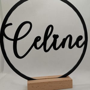 Könnte beinhalten: Schwarzer Metallkreis mit dem Namen "Celine" in kursivschrift. Der Kreis ist auf einem Holzsockel montiert.