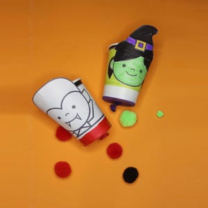 Puede incluir: Dos lanzaconfeti cilíndricos con diseños de personajes. Uno muestra un vampiro blanco con colmillos y base roja, mientras que el otro presenta una bruja de cara verde con sombrero negro y banda morada. Varios pompones pequeños rojos, negros y verdes.