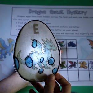 以下が含まれることがあります： 水色のドラゴンのイラスト、文字「E」、濃い青色の斑点が描かれた半透明の卵型の切り抜き。この切り抜きは、「Dragon Quest Mystery」というタイトルのアクティビティシート（グリッドとドラゴンの画像が印刷されている）にかざされています。