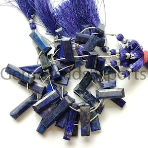 Puede incluir: Una colección de cuentas rectangulares facetadas de lapislázuli de varios tamaños. Las piedras azul oscuro están ensartadas con alambre plateado y van acompañadas de una borla azul oscuro.