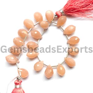 May include: A strand of faceted, pear-shaped peach-colored gemstones arranged in a circular pattern. The beads are strung on a thin silver-toned wire, with a vibrant coral-pink tassel attached to each end of the strand for a decorative finish.
