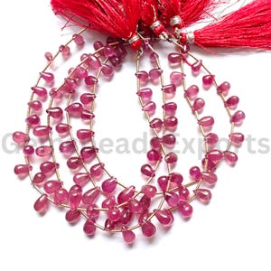 Puede incluir: Múltiples hilos de piedras preciosas de turmalina rosa, facetadas y con forma de lágrima, ensartadas en un hilo fino. Los hilos están unidos en la parte superior y asegurados con borlas decorativas de color rojo brillante y tono rosado.
