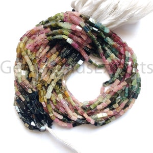 Op de afbeelding: Strengen veelkleurige toermalijn kralen. De kralen zijn rechthoekig en hebben een mix van roze, groene en zwarte tinten. De kralen zijn geregen met een witte kwast aan de bovenkant.