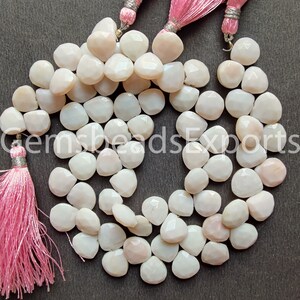 May include: A collection of faceted, off-white gemstone beads. The beads are in a teardrop shape and are strung together. Pink tassels are visible at the top and bottom of the image. The beads have a subtle, pearlescent sheen.