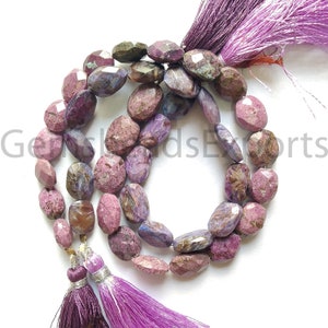 Puede incluir: Un collar de cuentas facetadas de piedra preciosa de charoíta en tonos morados, rosas y marrones. Las cuentas de forma ovalada están ensartadas, con una borla morada en cada extremo. Las piedras preciosas tienen una superficie pulida y reflectante.