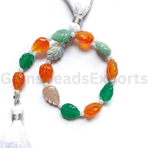 May include: A strand of gemstone beads in leaf shapes, featuring orange, green, and gray colors. The beads are interspersed with small white pearls. The strand is finished with a white tassel and silver-toned accents. The text "GemsBeadsExports" is visible.