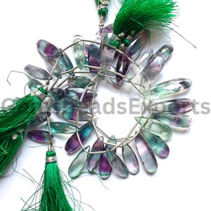 Puede incluir: Hebras de cuentas de piedras preciosas de fluorita pulida en tonos morado, verde y transparente. Las cuentas están ensartadas con alambre de plata y acentuadas con borlas verdes. Las piedras preciosas tienen forma de lágrima.