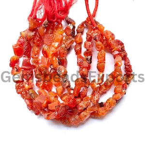 May include: A strand of irregular, tumbled carnelian gemstone beads in shades of orange and red, strung on a red cord with a red tassel at the top. The text Gemsbead Exports is overlaid across the centre of the gemstone bead arrangement.
